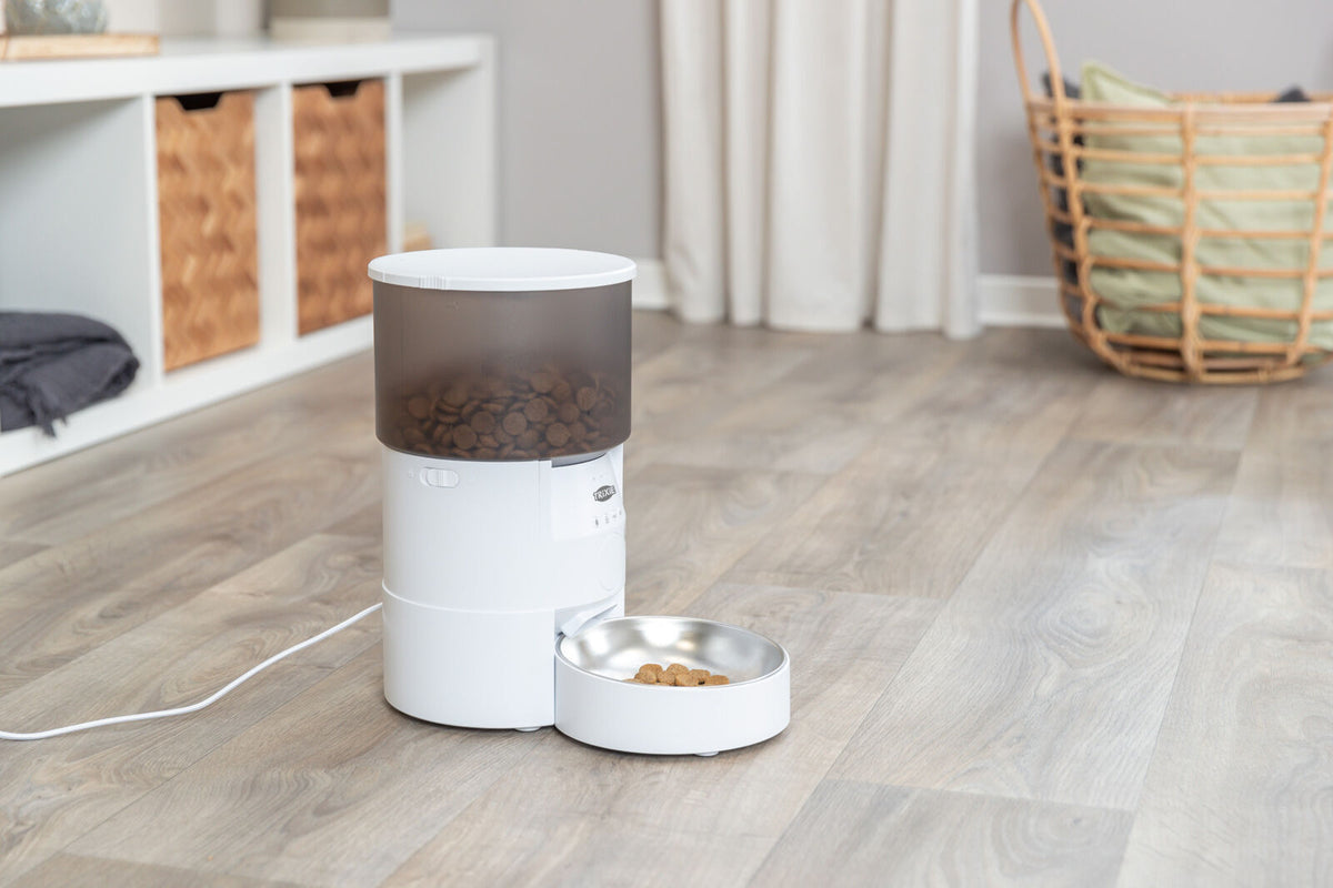 Ein automatischer Futterspender mit durchscheinendem Behälter steht auf einem Holzboden neben einem Körbchen.