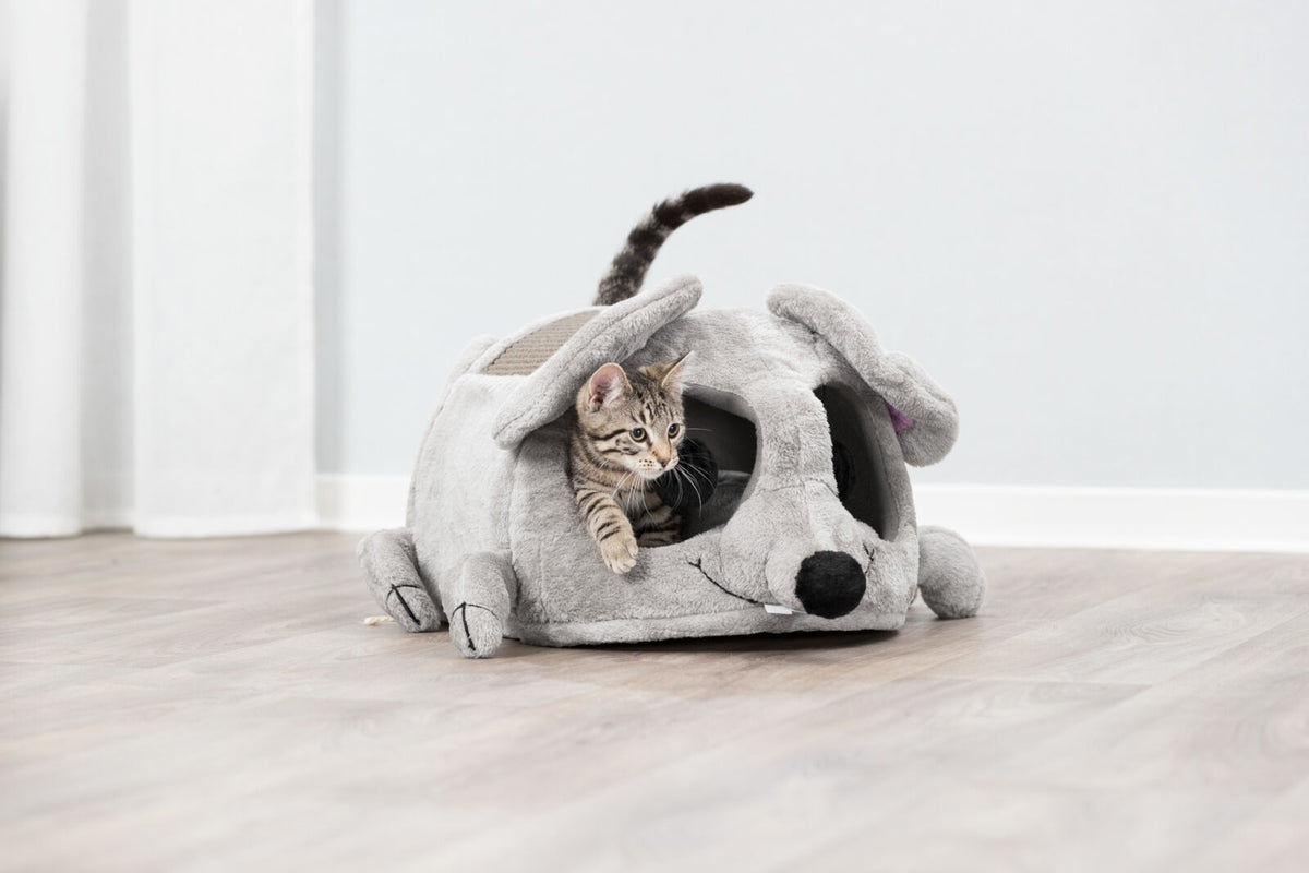 Kleine Katze schaut aus einer grauen Plüschhöhle in Form einer Maus auf hellem Holzboden hervor.