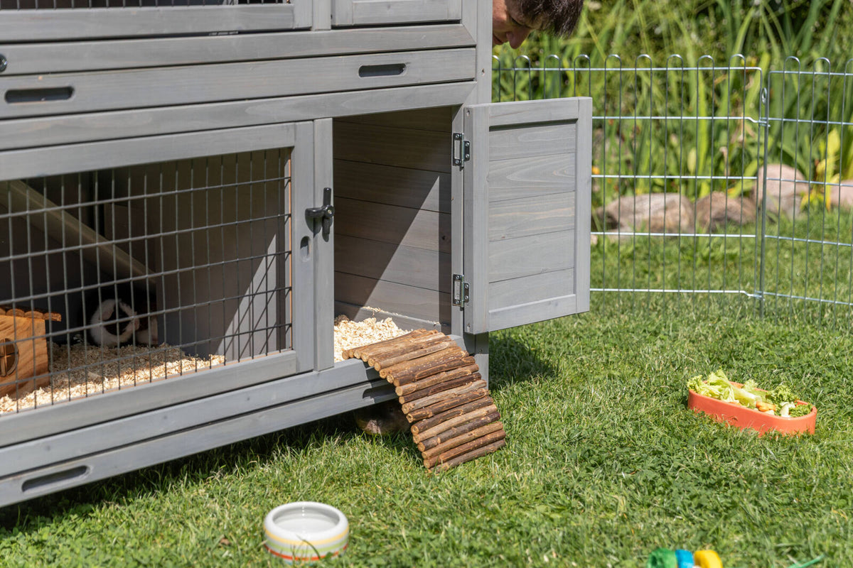 Graues Kleintiergehege mit offener Tür steht auf Gras, neben einem Zaun, Futternapf und Spielzeug.