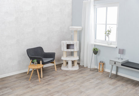 Wohnzimmer mit grauem Sessel, Holztisch, Kratzbaum und Fenster mit weißem Vorhang, Pflanzen auf Boden und Tisch.