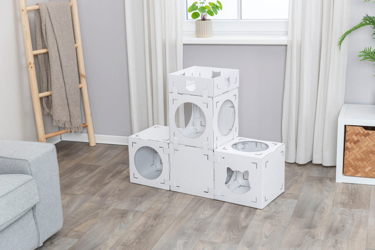 Weiße modulare Würfel mit kreisförmigen Öffnungen stehen in einem Zimmer auf einem Holzfußboden, im Hintergrund Pflanzen und ein graues Sofa.