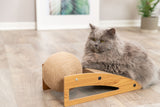 Graue Katze neben einem hölzernen Kratzbrett mit einer großen, kugelförmigen Sisalrolle in einem hellen Zimmer auf Holzboden.