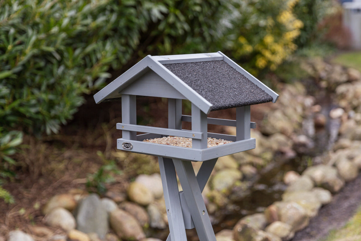 Graues hölzernes Vogelhaus mit schwarzem Dach auf einem Ständer, im Garten positioniert, mit Hintergrund aus Büschen und Steinen.