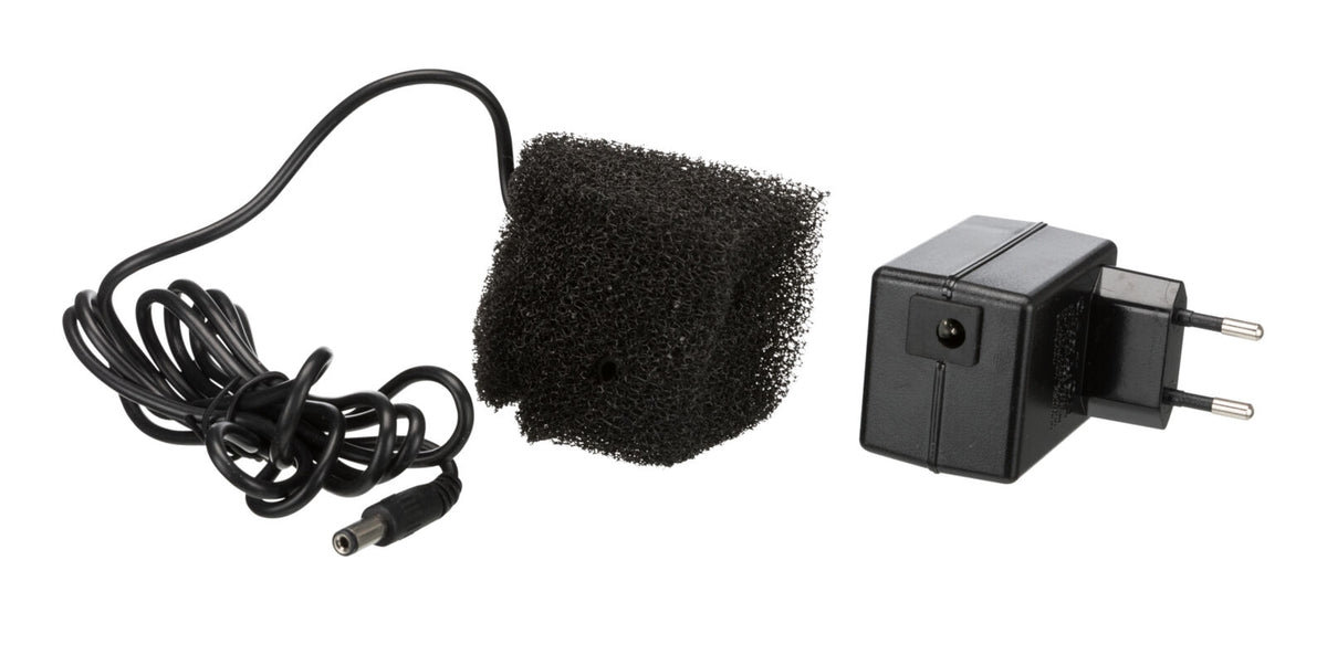 Ein schwarzes Netzteil mit europäischem Stecker neben einem schwarzen Schaumstoffstück und einem Kabel.