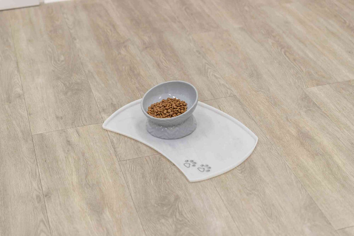 Graue Futterschale mit Trockenfutter auf weißer Matte mit Pfotensymbolen auf hellem Holzboden.