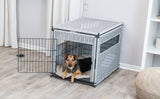 Ein kleiner brauner Hund liegt in einem grauen, geflochtenen Tierkäfig mit offener Tür auf einem beigen Kissen.