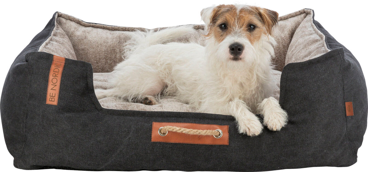 Ein Hund liegt in einem grauen Hundebett mit beigem Innenfutter, darauf ein Lederetikett mit Aufschrift "BE NORDIC".