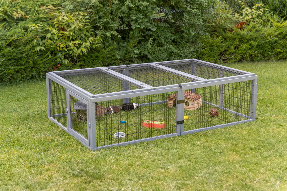 Freigehege aus Metallgitter steht auf einer Wiese, mit Kleintierunterkunft und Spielzeug.