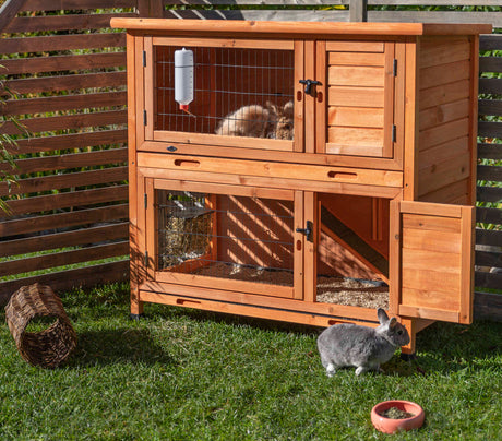 Holzkäfig mit zwei Etagen in einem Garten, oben ein Kaninchen und ein weiteres am Boden; Trinkflasche und Futternapf sichtbar.