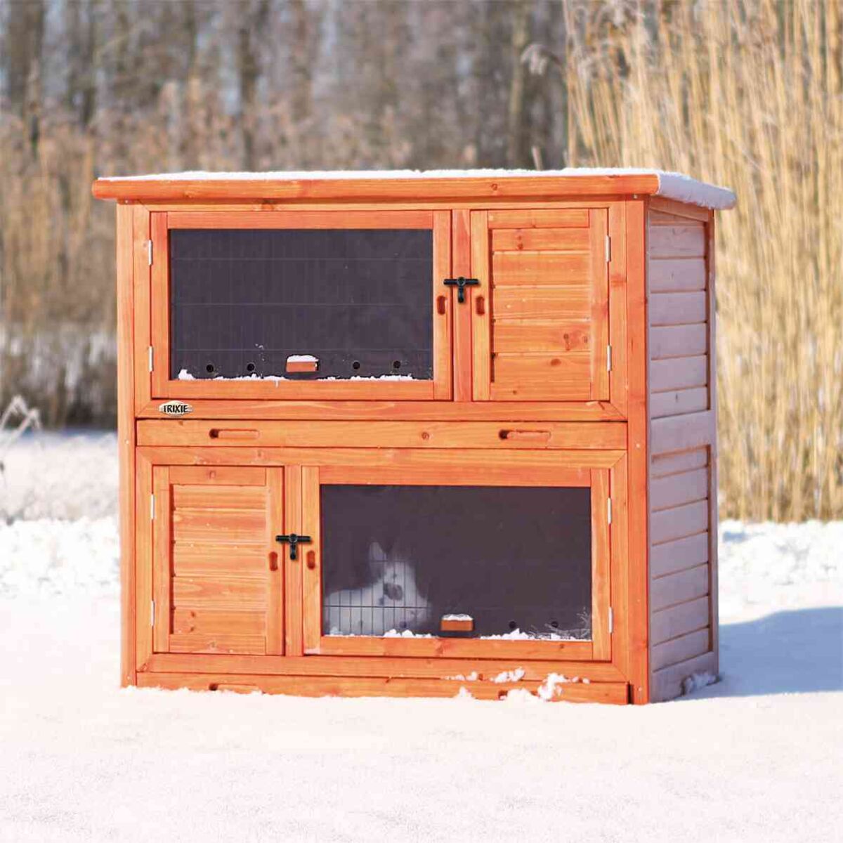 Holzstall mit zwei Ebenen, Gittertüren und Schnee bedeckt, im Inneren ist ein schwarzes und weißes Tier sichtbar.