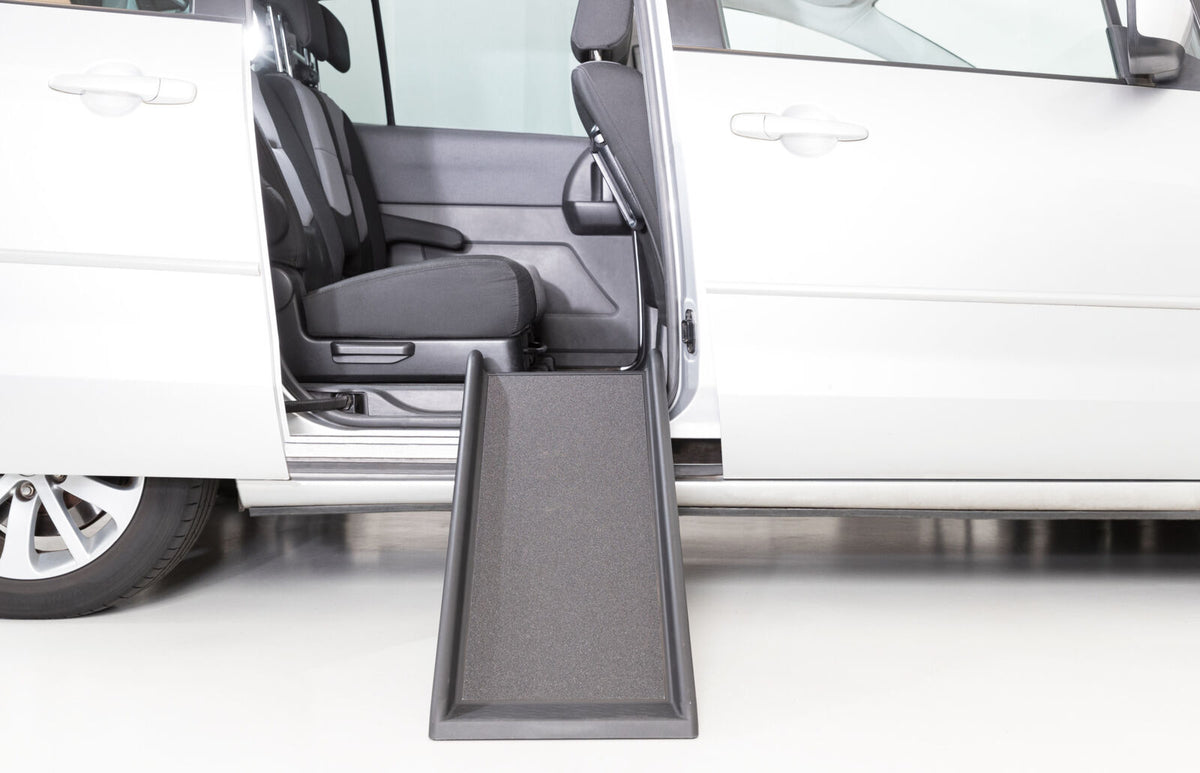 Silbernes Auto mit geöffneter Tür, schwarze Rampe führt von der Bordsteinkante zum Innenraum.